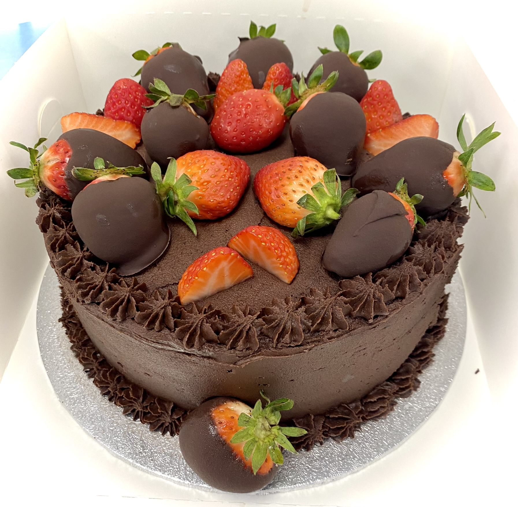 A cake topped with whole strawberries and chocolate-covered strawberries. The cake has an outer layer of chocolate frosting with decorative chocolate piped frosting around the edges. It sits on a silver cake board inside a white box.